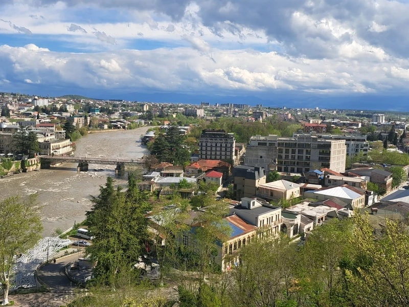 Vista panorámica de Kutaisi con el río Rioni cruzado por un puente, edificios variados y vegetación verde bajo un cielo nublado