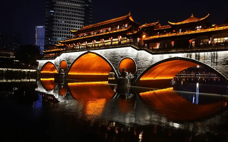 Puente tradicional chino iluminado en tonos rojos sobre un río nocturno, con arcos reflejados en el agua y rascacielos al fondo
