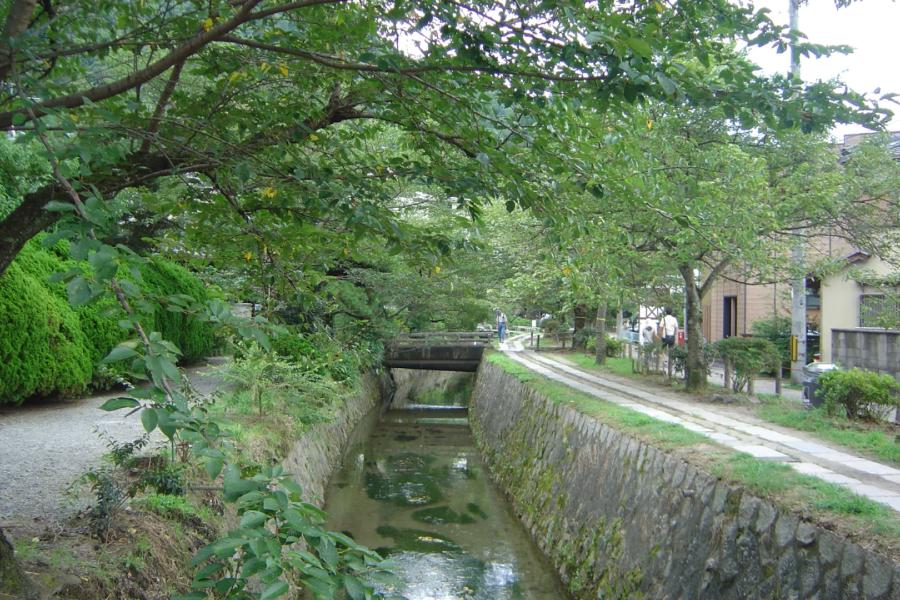 Un canal sereno en Kioto con vegetación exuberante, puente peatonal y casas tradicionales al fondo