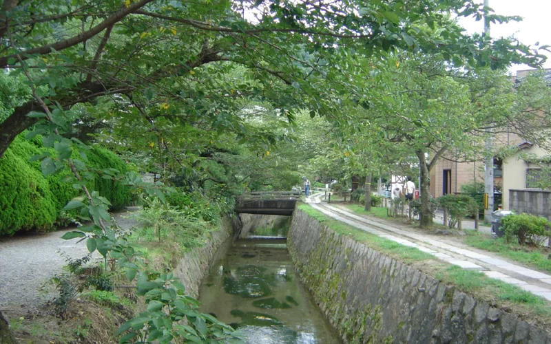 Un canal sereno en Kioto con vegetación exuberante, puente peatonal y casas tradicionales al fondo