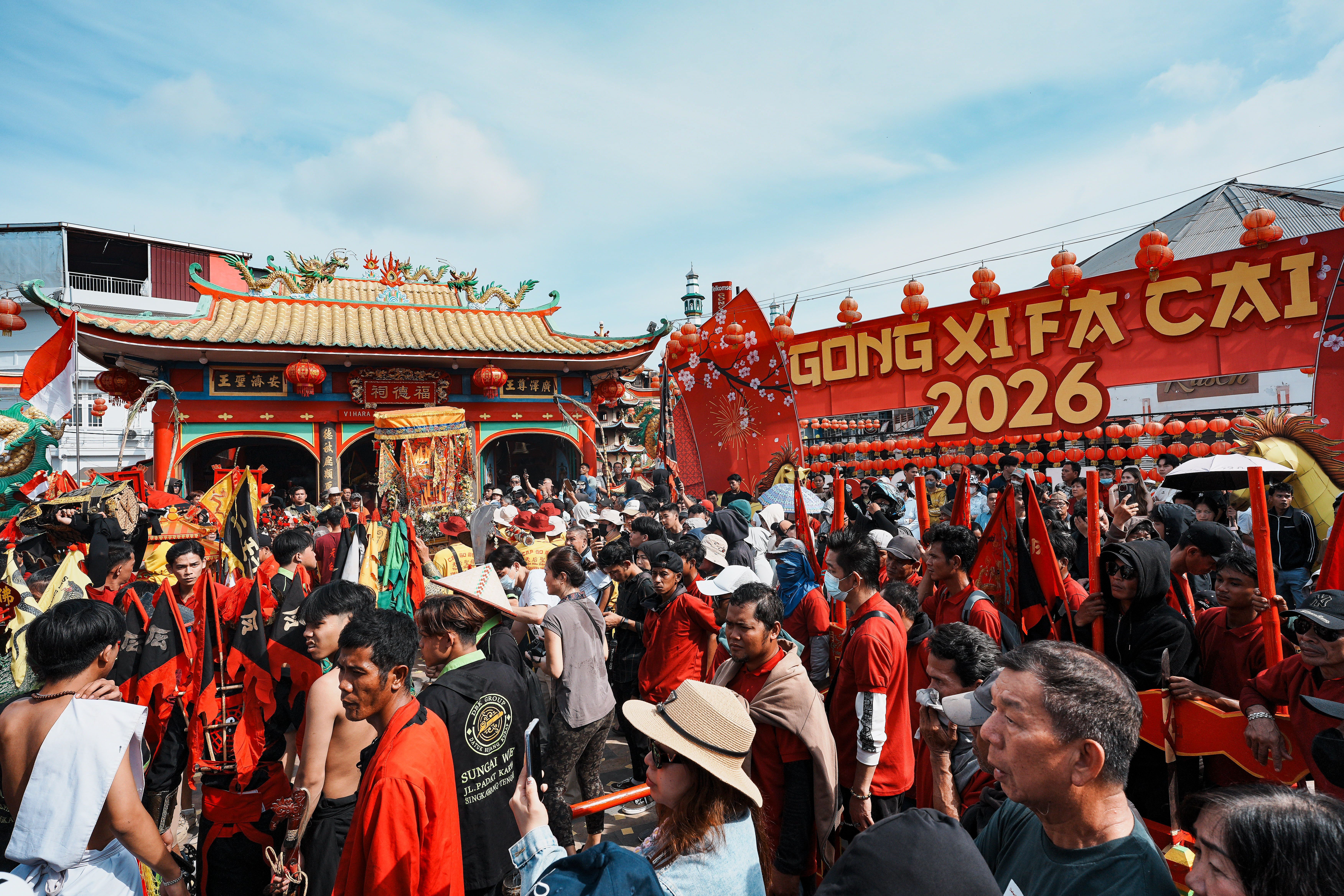 Multitud de personas en una celebración callejera con un templo chino y una pancarta roja.