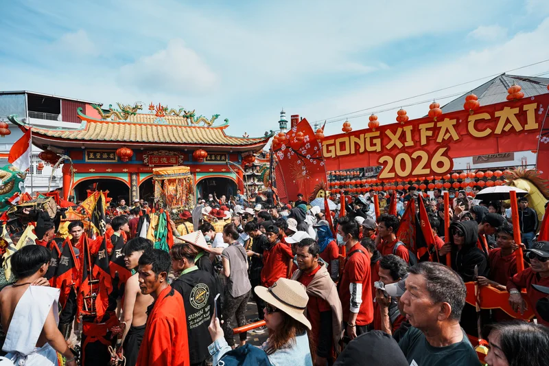 Multitud de personas en una celebración callejera con un templo chino y una pancarta roja.