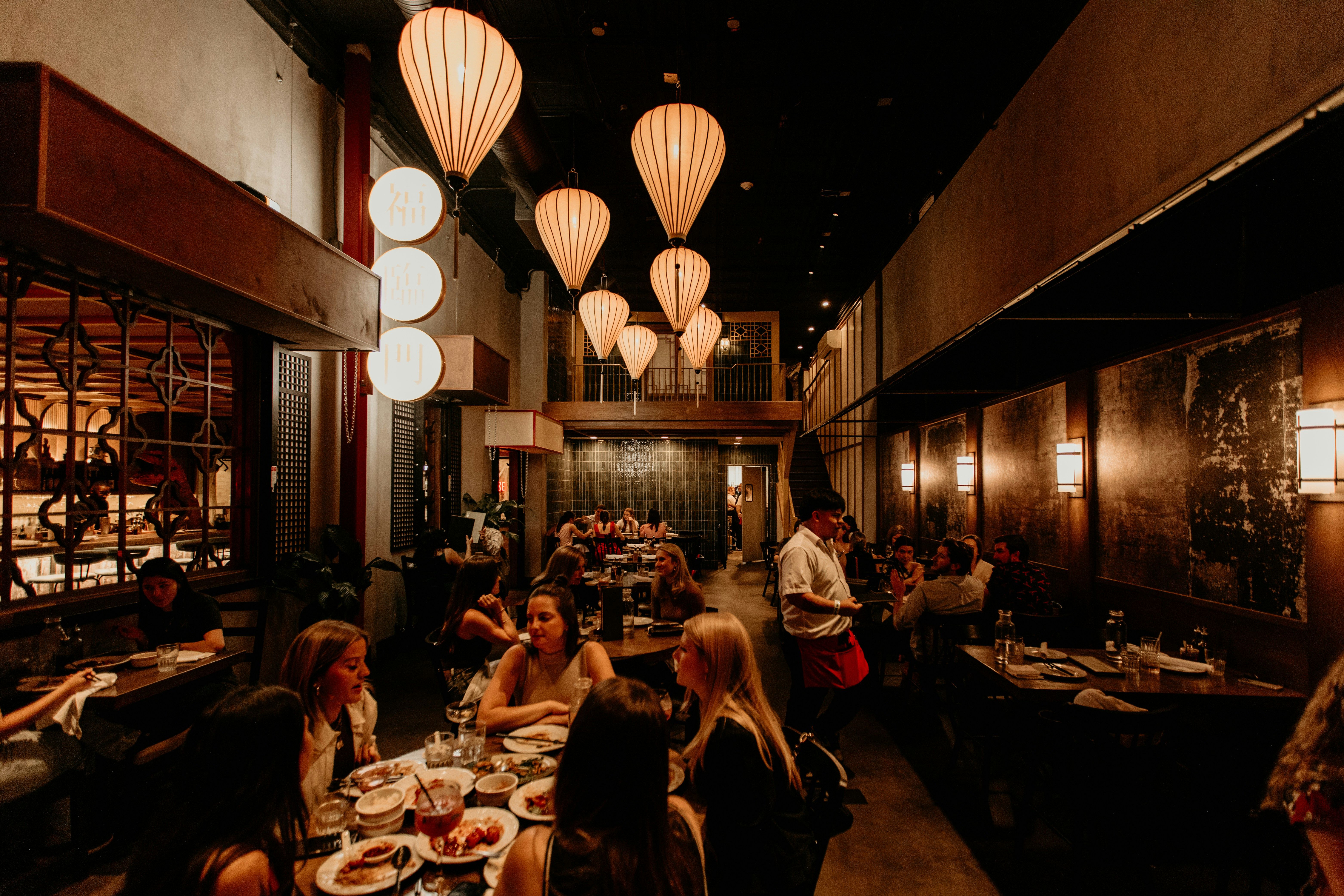 Interior de un restaurante moderno con lámparas colgantes, mesas llenas de comensales y un camarero.