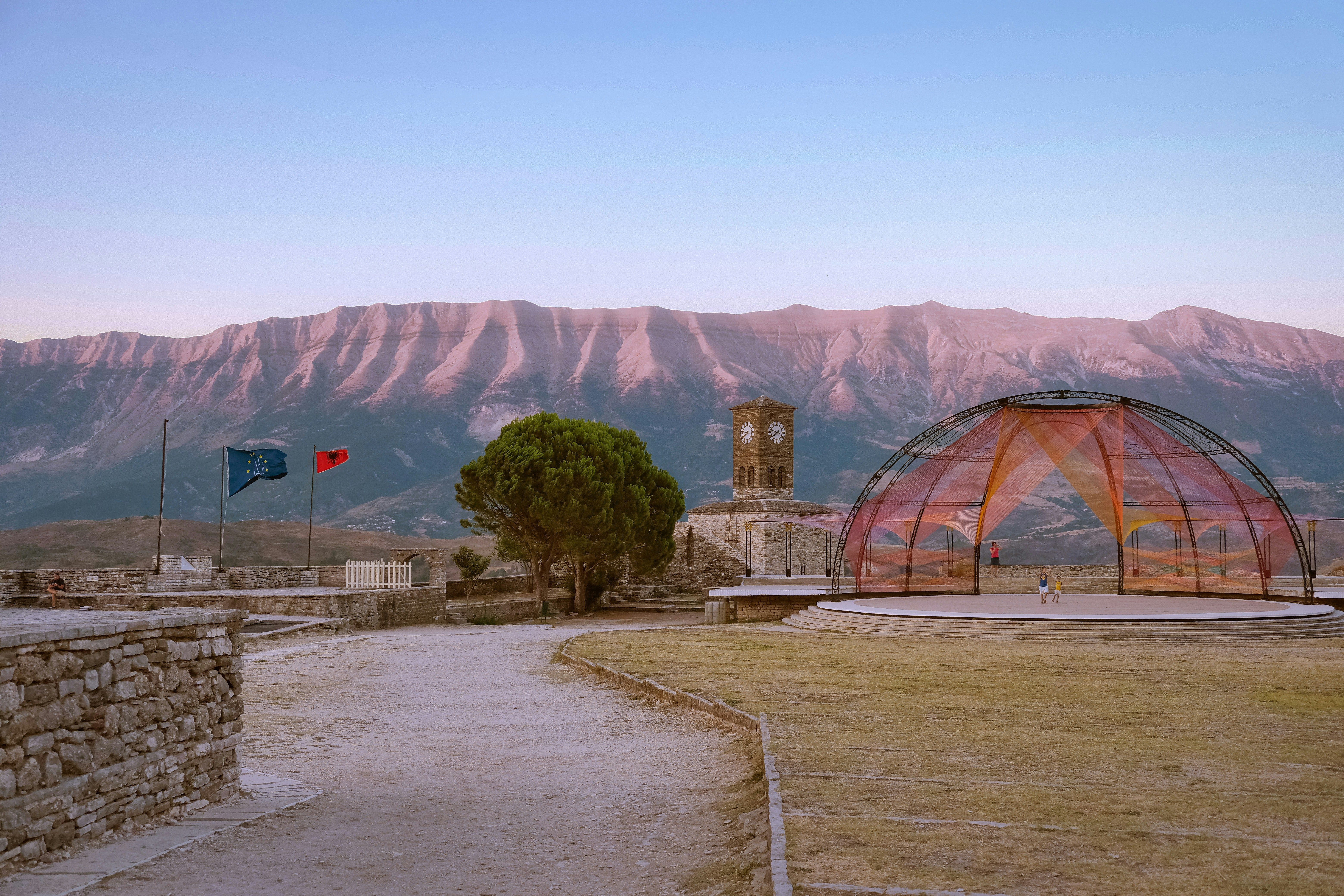 Vista de un castillo con torre del reloj, banderas, un escenario moderno y montañas al fondo.
