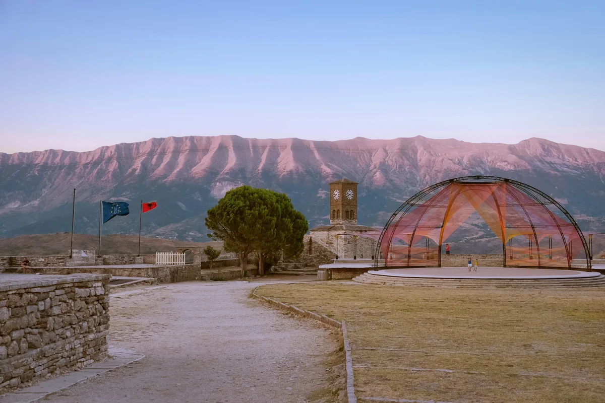 Vista de un castillo con torre del reloj, banderas, un escenario moderno y montañas al fondo.