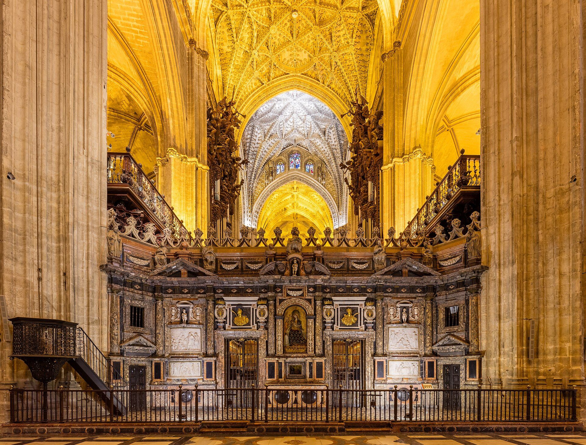 Interior de una catedral con el trascoro ricamente decorado y techos abovedados iluminados en tonos dorados.