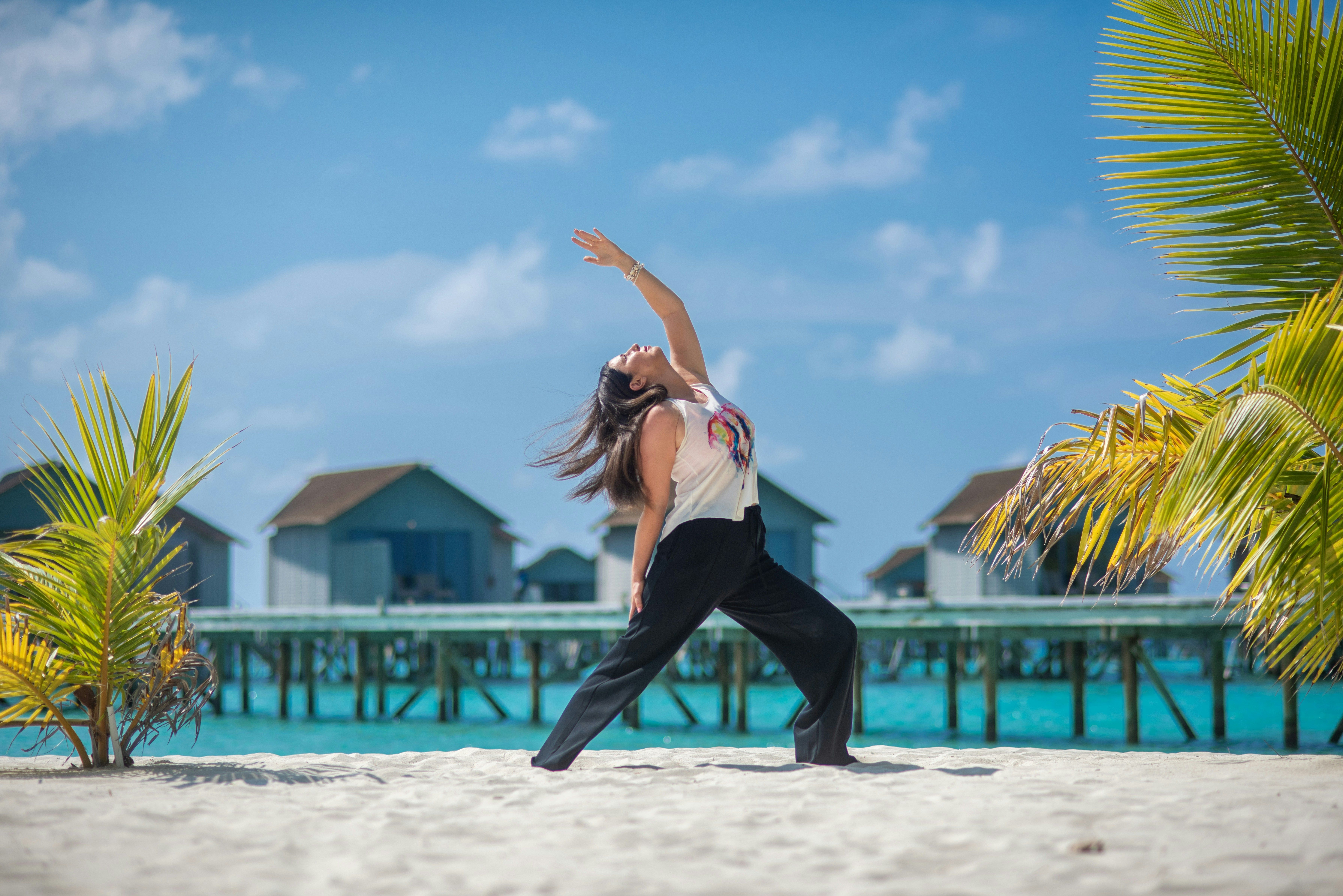Mujer haciendo yoga en la playa, con cabañas sobre el agua y palmeras de fondo.