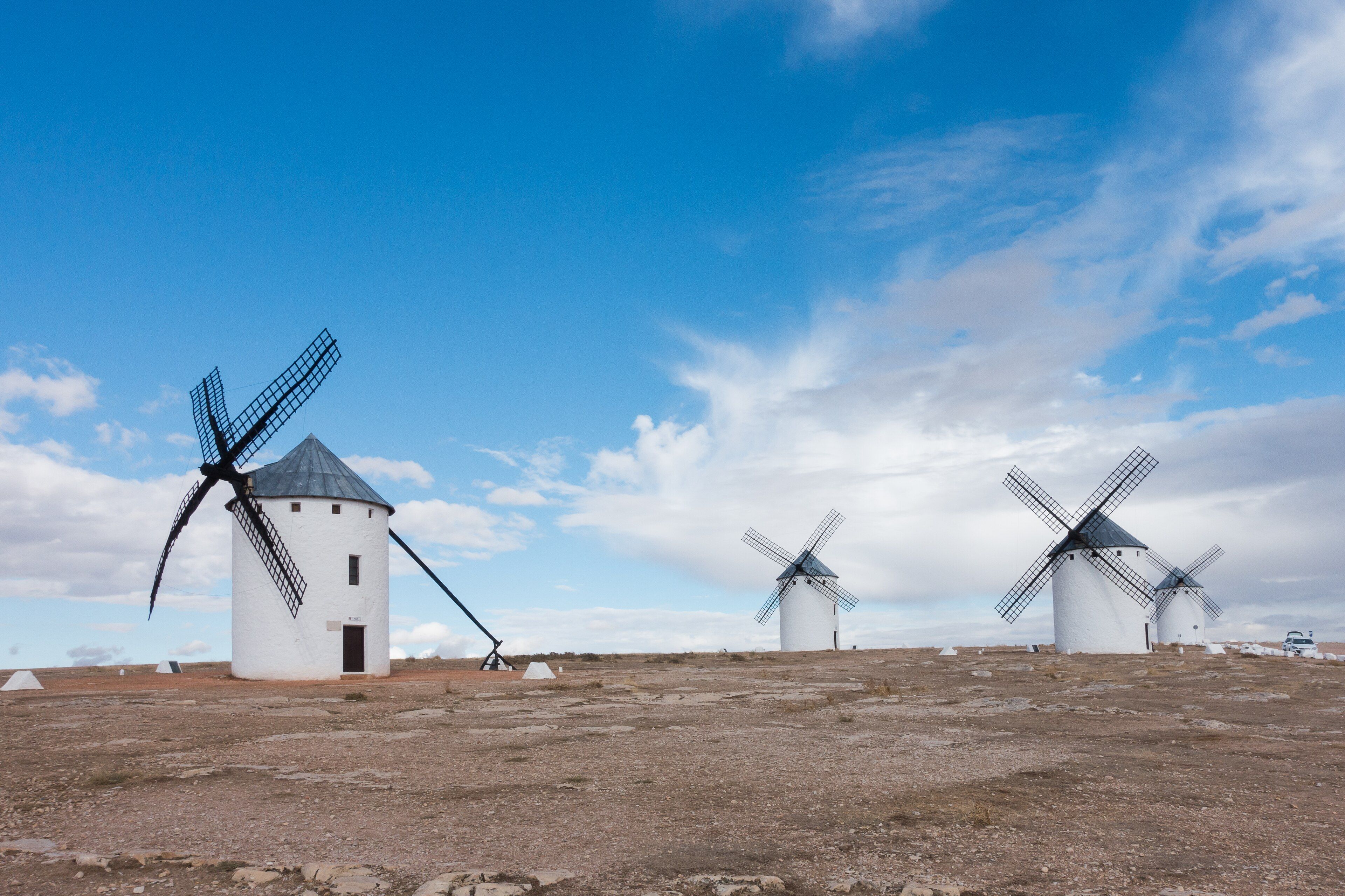 Cuatro molinos de viento blancos con tejado cónico gris y aspas negras sobre un cerro pedregoso, bajo un cielo azul con nubes.