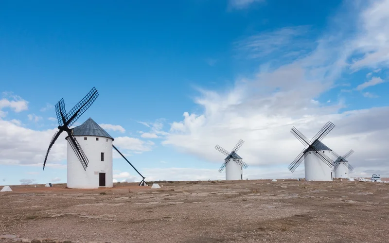 Cuatro molinos de viento blancos con tejado cónico gris y aspas negras sobre un cerro pedregoso, bajo un cielo azul con nubes.