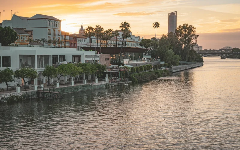 Vista del río Guadalquivir al atardecer, con edificios ribereños, palmeras y un puente a lo lejos.