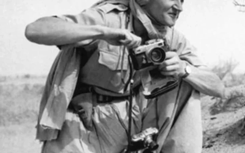 Fotógrafo con turbante y chaleco caqui arrodillado en dunas desérticas, ajustando su cámara antigua en blanco y negro