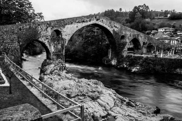Puente medieval de piedra con arcos sobre un río rocoso, en blanco y negro.