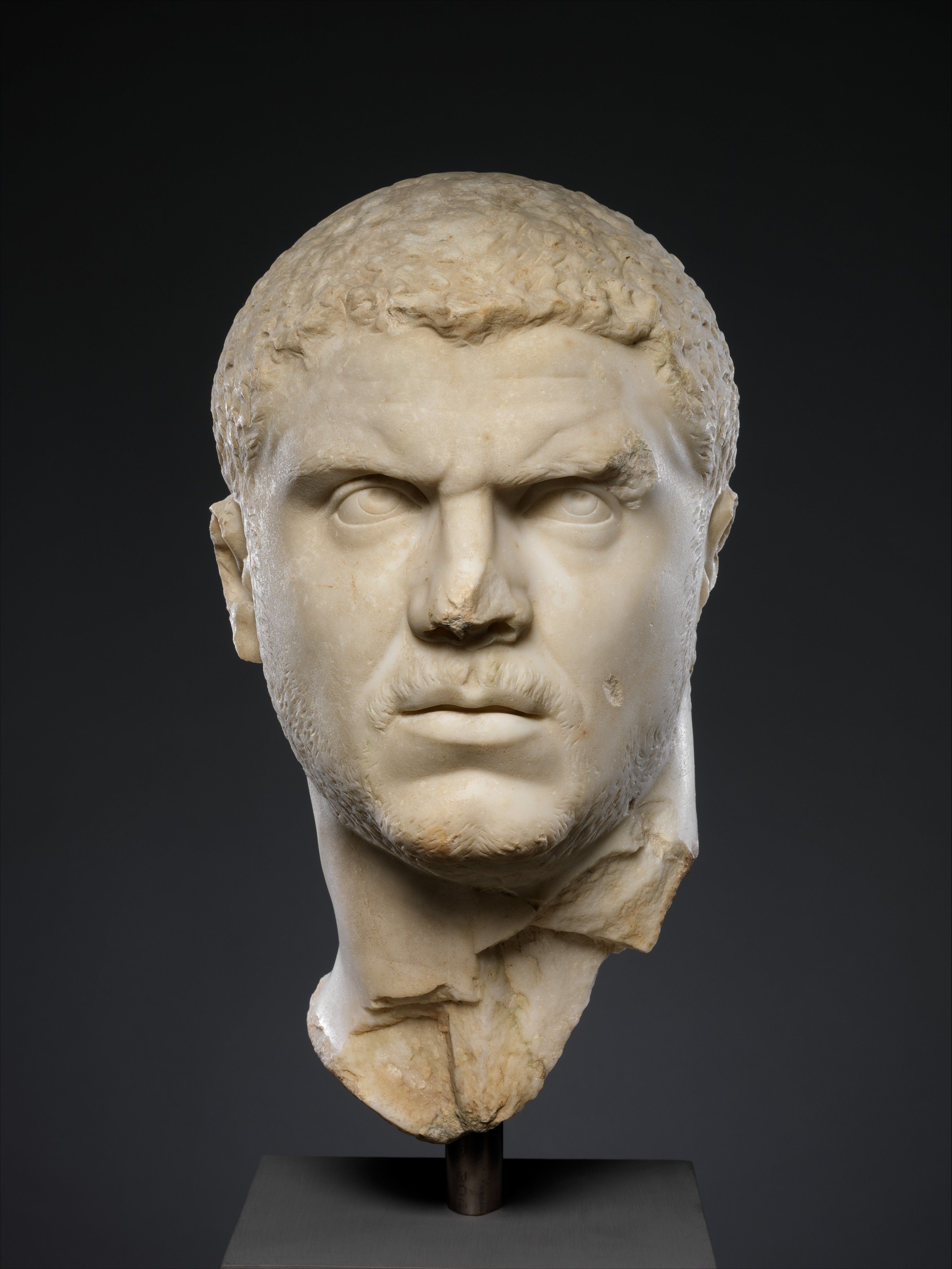 Busto de mármol de un hombre romano con cabello corto y barba incipiente, sobre un pedestal oscuro.