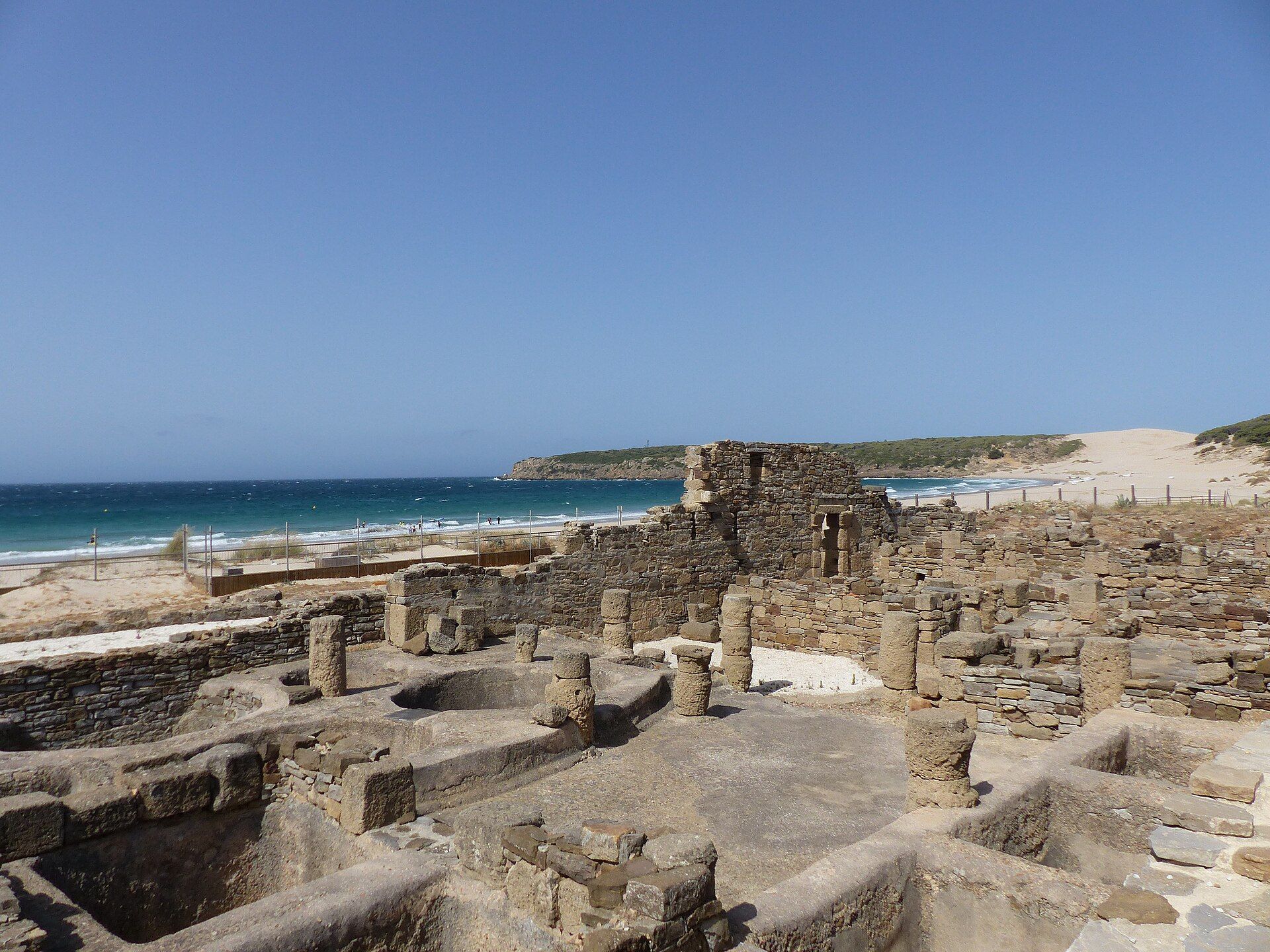 Ruinas de piedra de una antigua factoría de garum junto a una playa y el mar bajo un cielo azul claro.