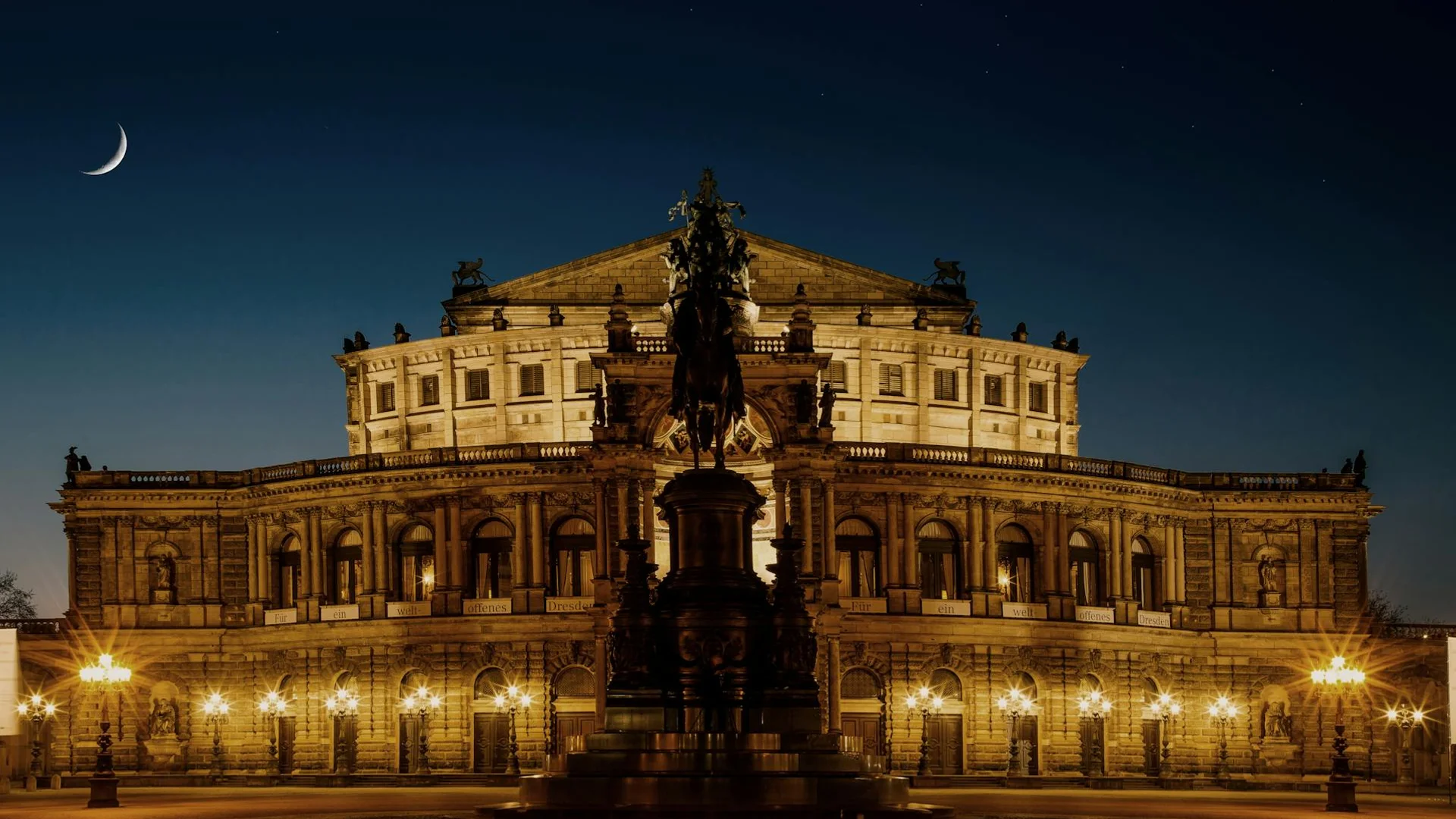 Vista nocturna de la Semperoper en Dresden, con fachada barroca iluminada, estatua central y luna creciente en el cielo