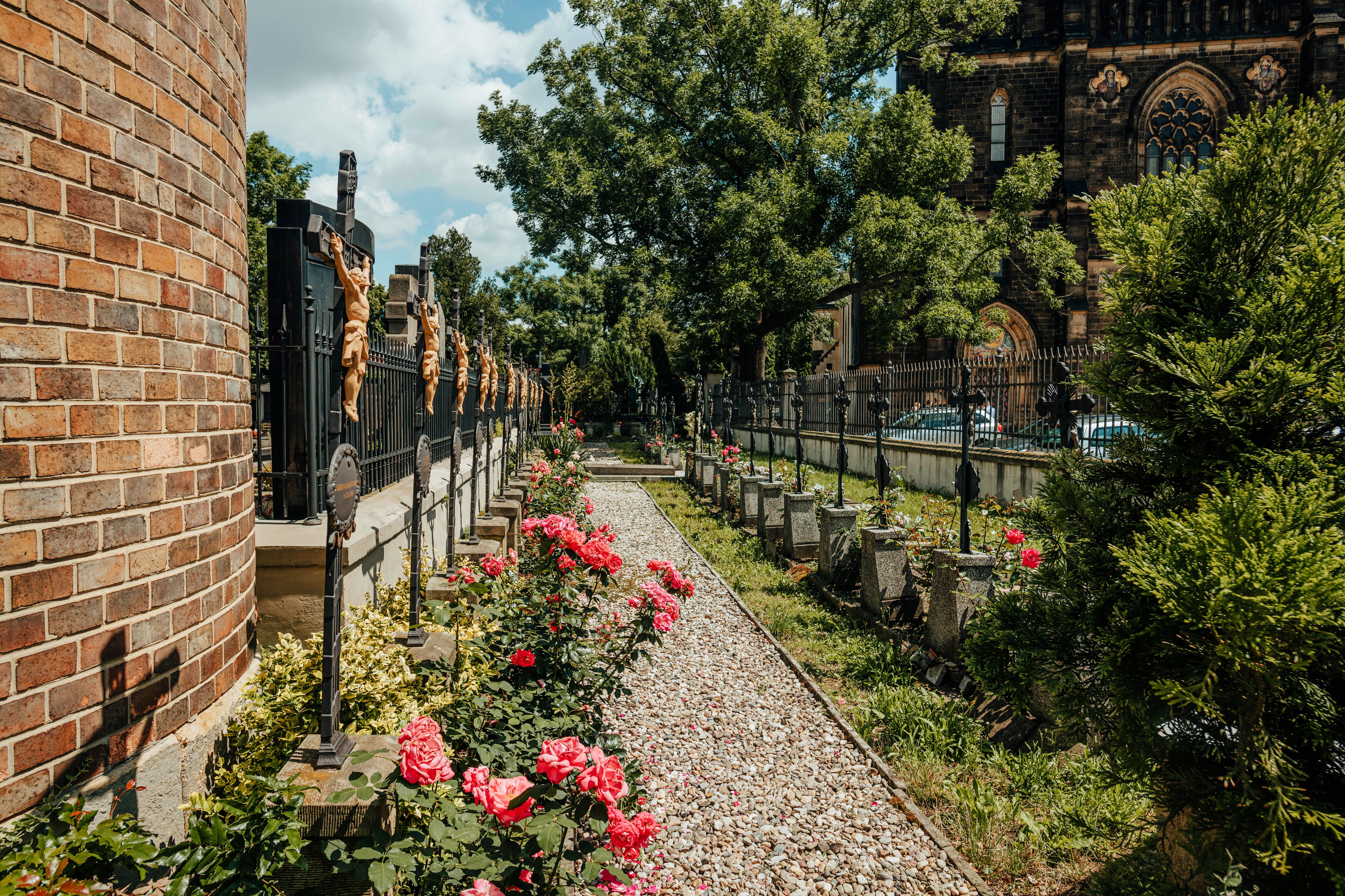 Camino de grava flanqueado por rosas rojas y cruces en un cementerio con un edificio de ladrillo.