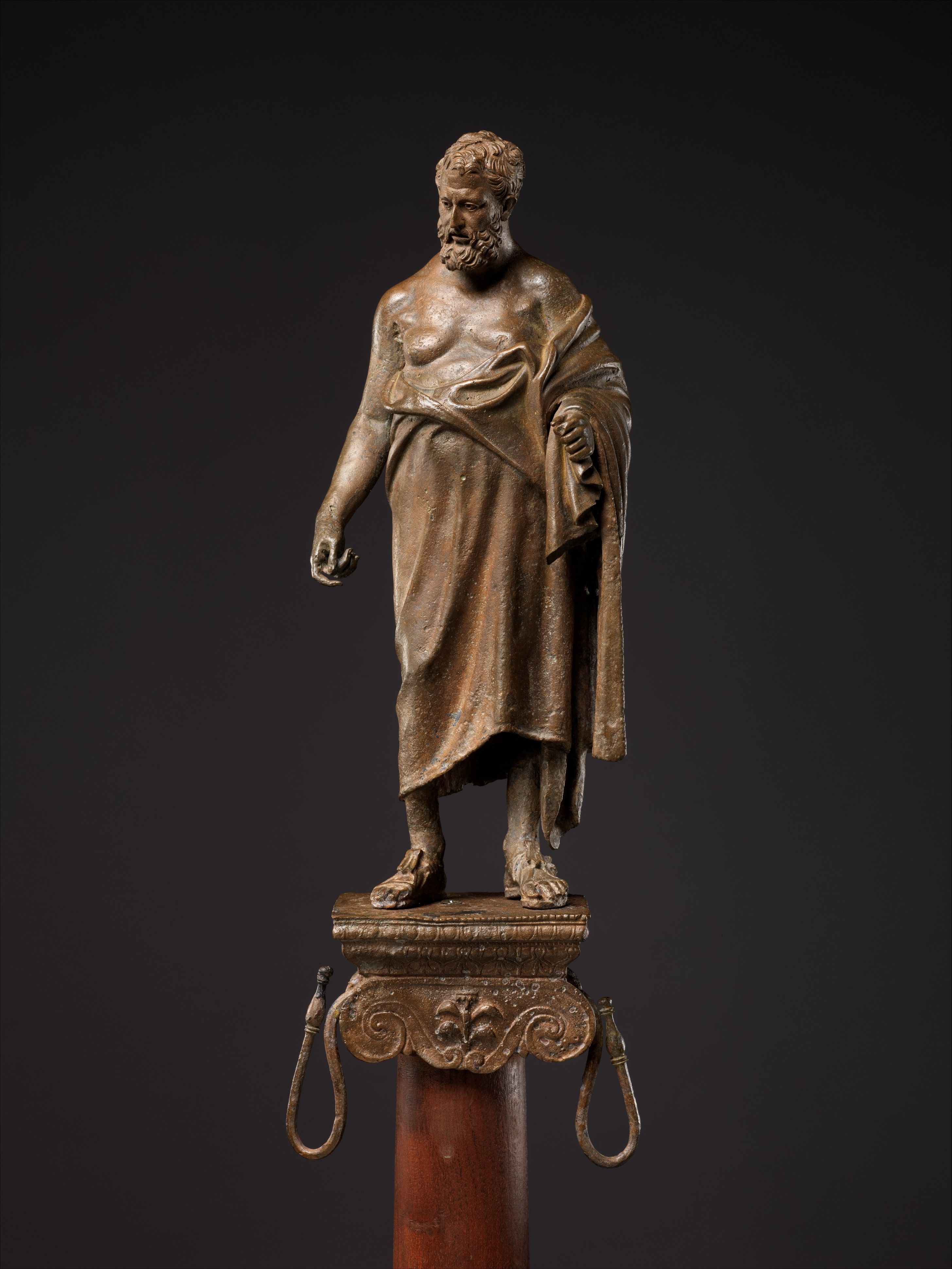 Estatua de bronce de un filósofo barbudo con túnica sobre un pedestal ornamentado de madera.