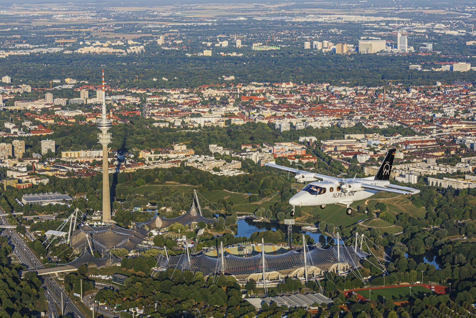 Vista aérea de Múnich con el Allianz Arena, el Olympiapark, la torre de televisión, un avión blanco volando y edificios rodeados de parques y lagos