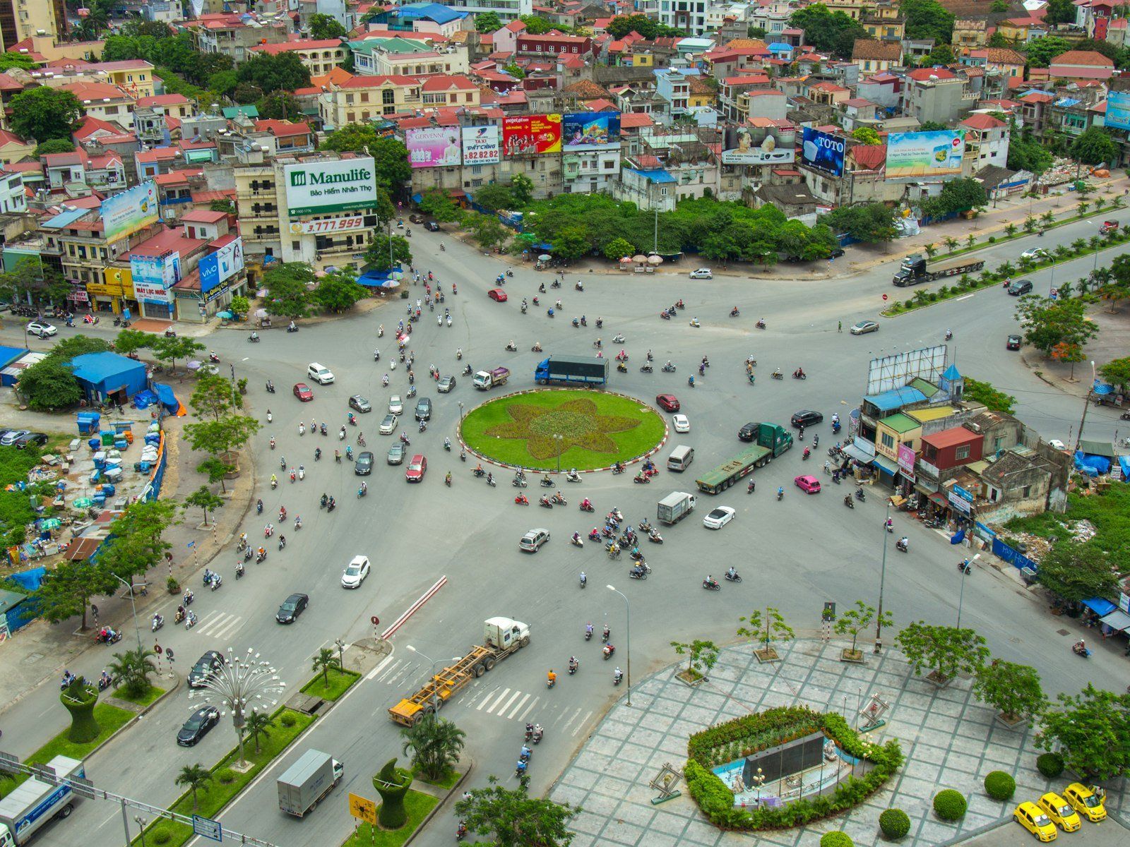 Vista aérea de una intersección bulliciosa en Hue, con motos y autos circulando alrededor de un jardín central, edificios de techos rojos y anuncios
