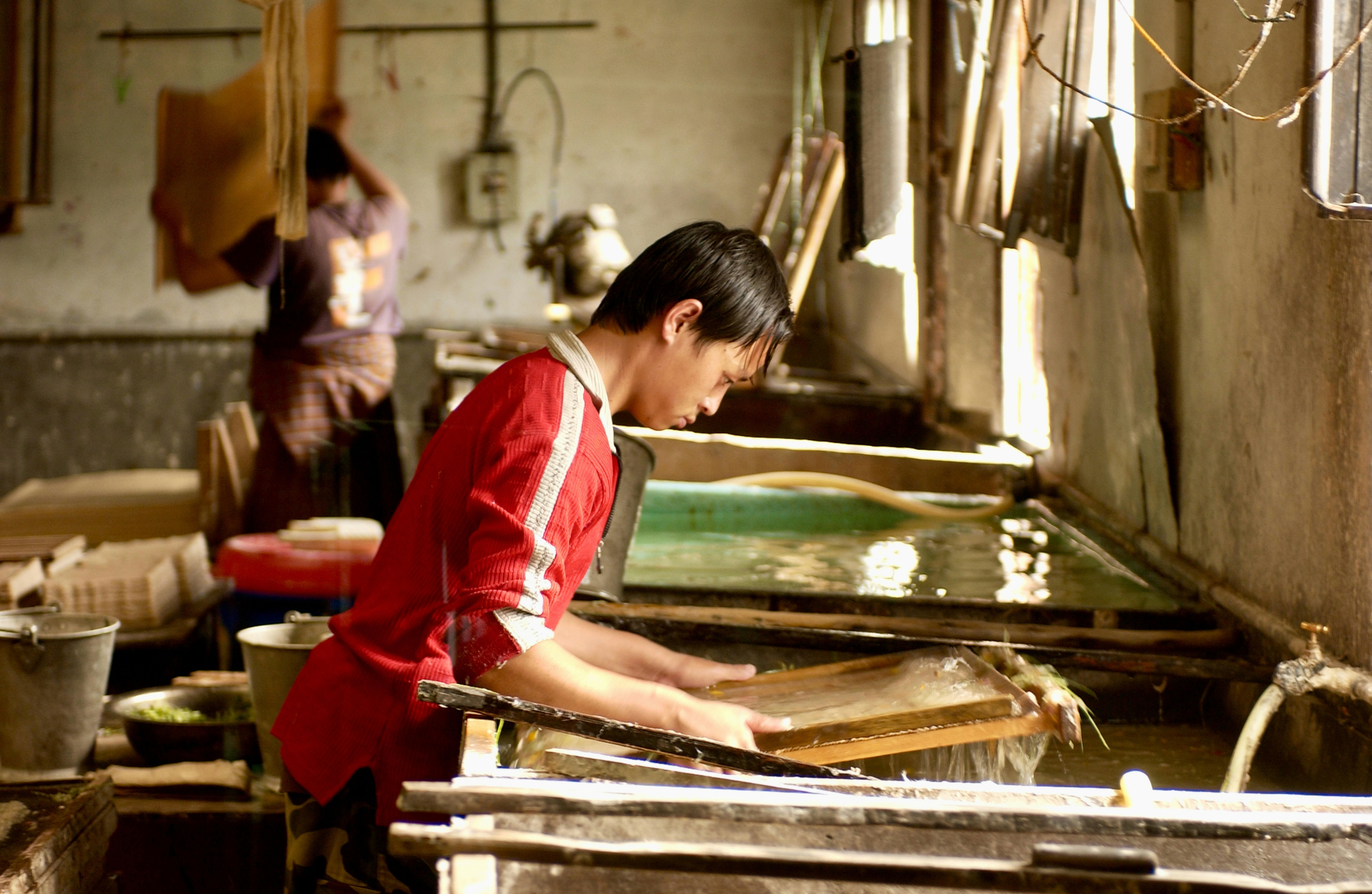 Hombre en camisa roja trabajando con una bandeja de malla en un taller de fabricación de papel.
