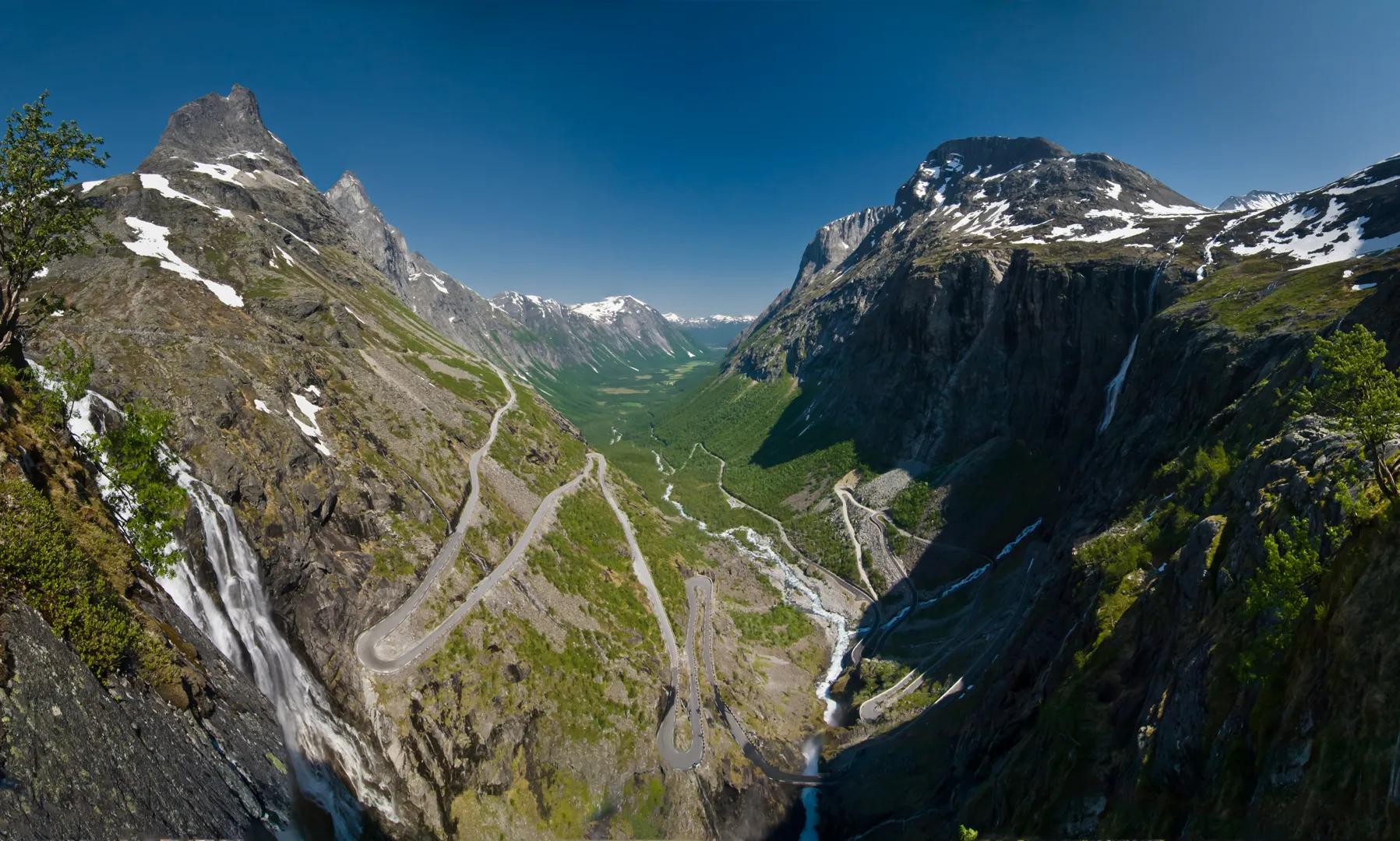Vista panorámica de un valle montañoso nevado en Noruega, con carretera sinuosa descendiendo entre verdes prados y cascadas bajo cielo azul