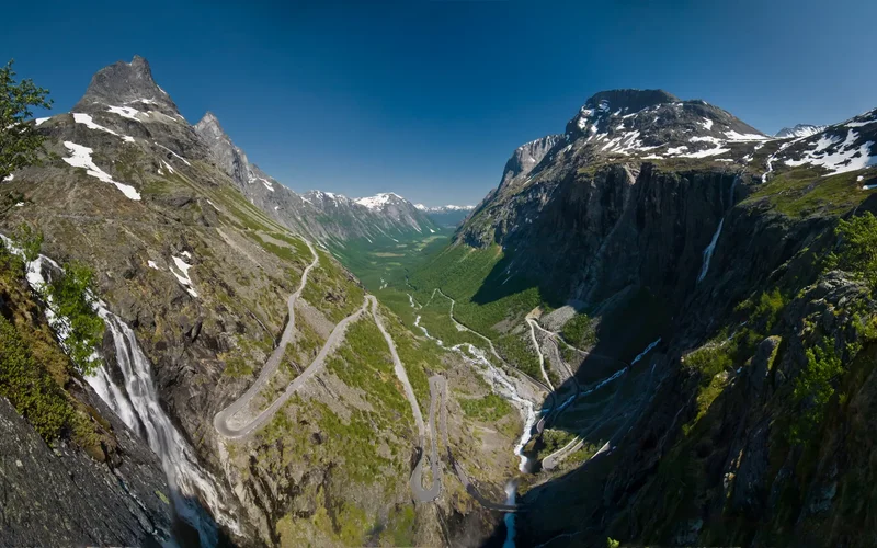 Vista panorámica de un valle montañoso nevado en Noruega, con carretera sinuosa descendiendo entre verdes prados y cascadas bajo cielo azul