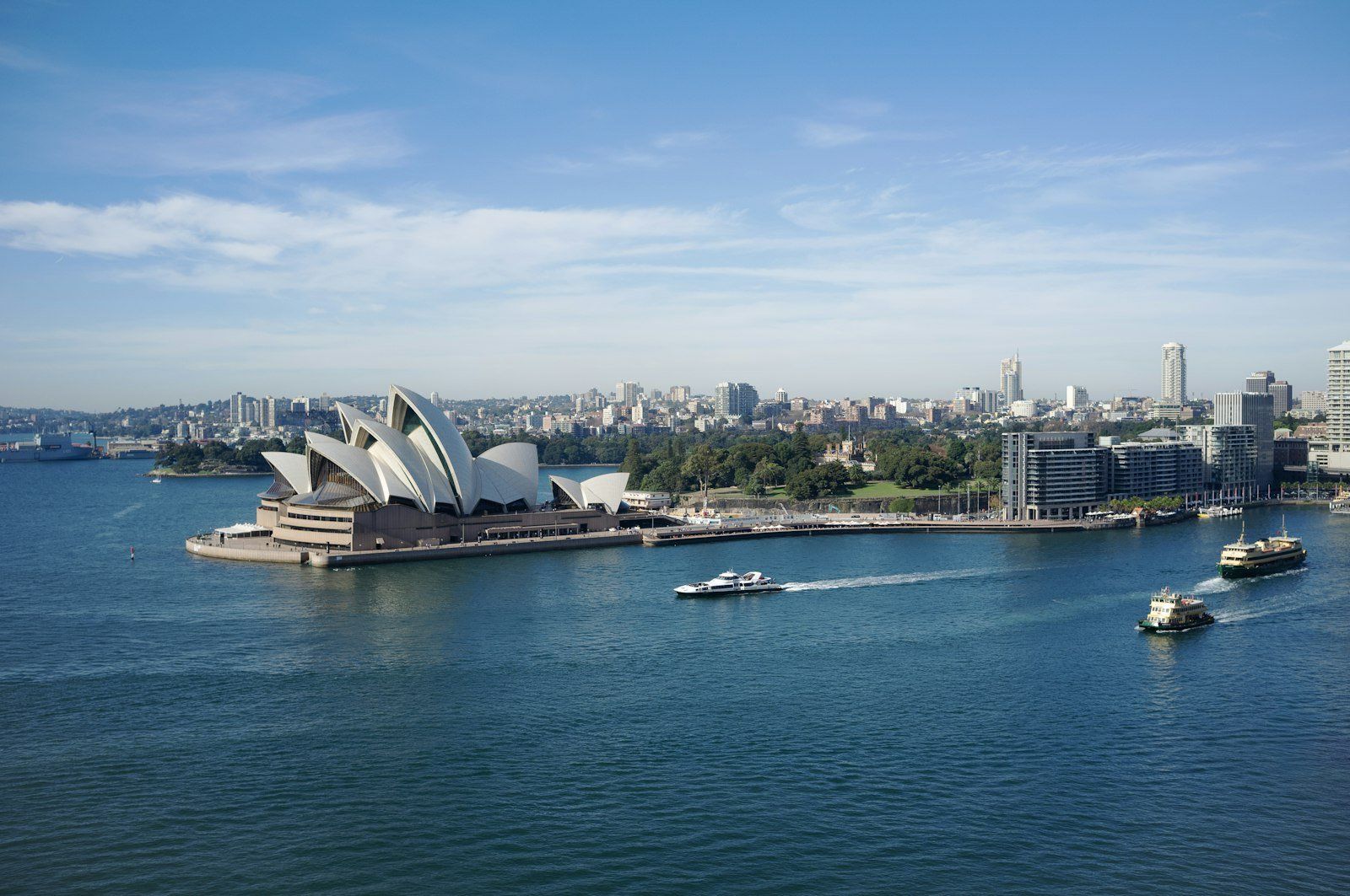 La Ópera de Sídney con su techo de velas junto al puerto, skyline de la ciudad al fondo, ferries en el agua azul y cielo con nubes