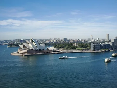 La Ópera de Sídney con su techo de velas junto al puerto, skyline de la ciudad al fondo, ferries en el agua azul y cielo con nubes