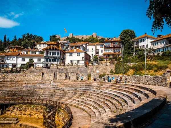 Vista panorámica de un antiguo anfiteatro de piedra con casas y una fortaleza en la colina al fondo.