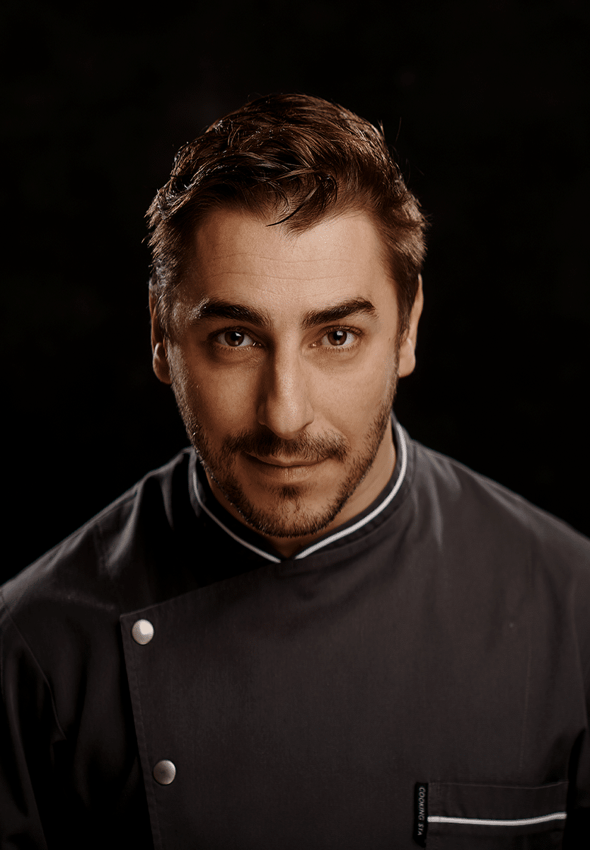 Retrato de Jordi Roca, chef con cabello castaño corto, ojos claros y barba corta, vistiendo chaqueta de chef gris oscura con cuello blanco, fondo