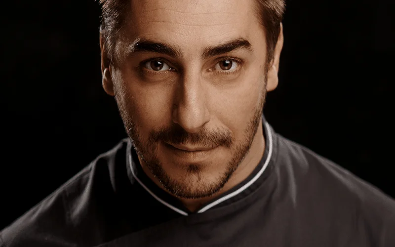 Retrato de Jordi Roca, chef con cabello castaño corto, ojos claros y barba corta, vistiendo chaqueta de chef gris oscura con cuello blanco, fondo