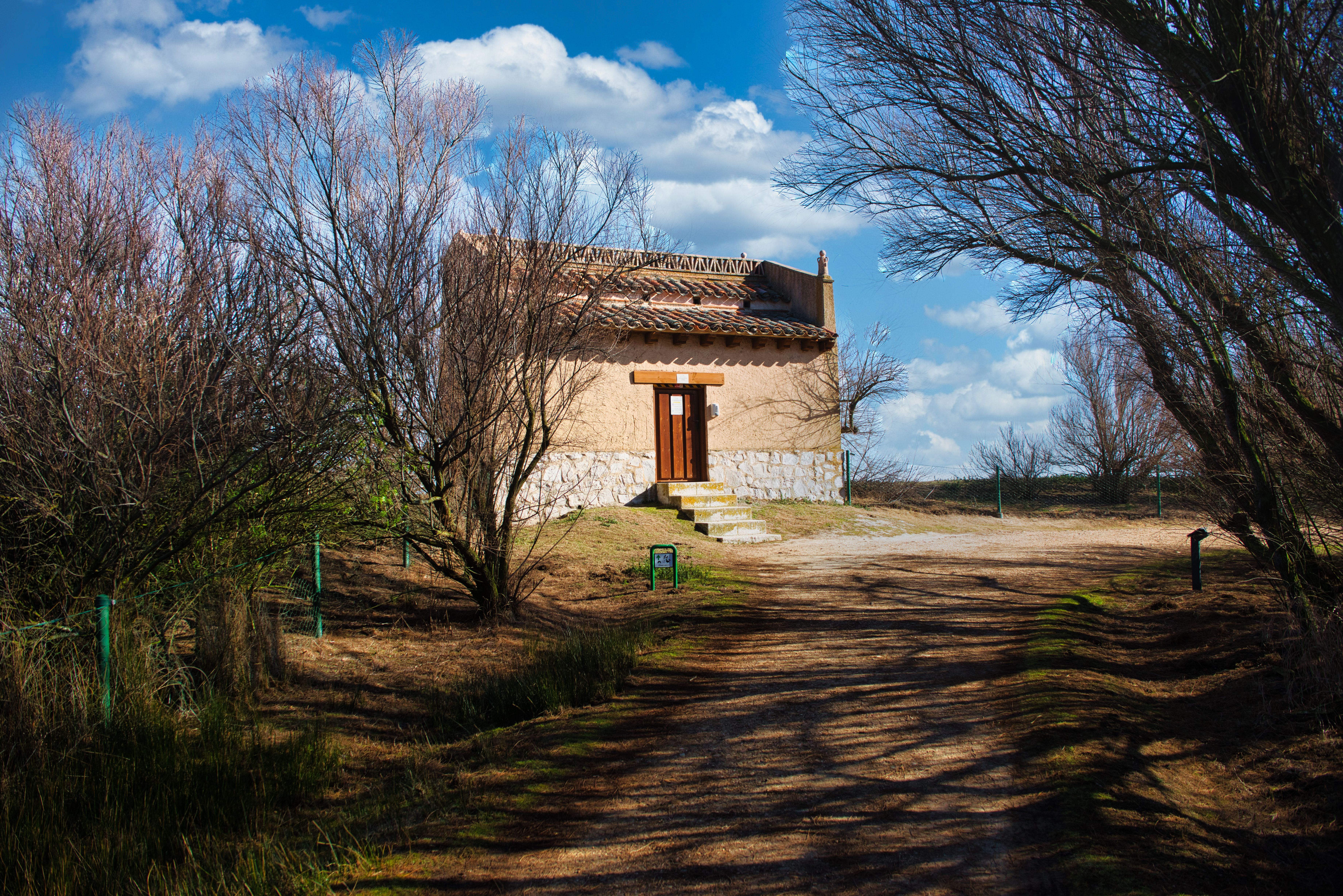 Edificio de piedra y adobe con tejado de tejas, rodeado de árboles sin hojas y cielo azul.
