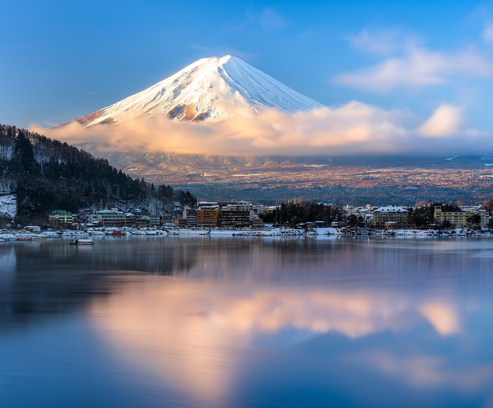 Monte Fuji nevado con nubes rosadas al amanecer, reflejado en un lago tranquilo con edificios en la orilla.