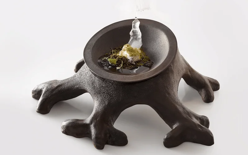 Postre en plato de tronco oscuro con raíces, espuma blanca, hierbas verdes y líquido negro simulando un bosque lluvioso