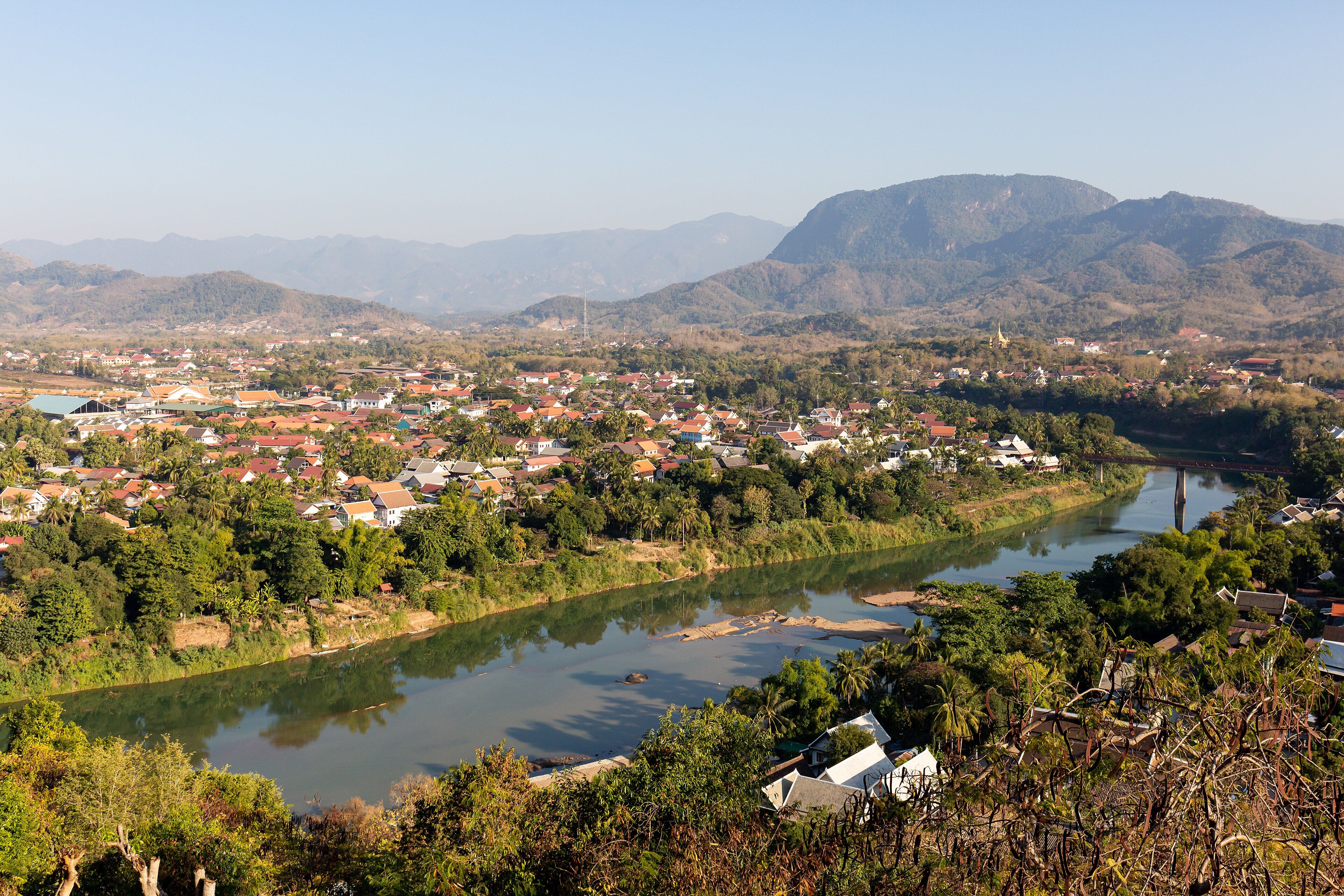 Vista desde lo alto de un río que serpentea entre casas de tejados rojos y palmeras, con montañas boscosas al fondo.