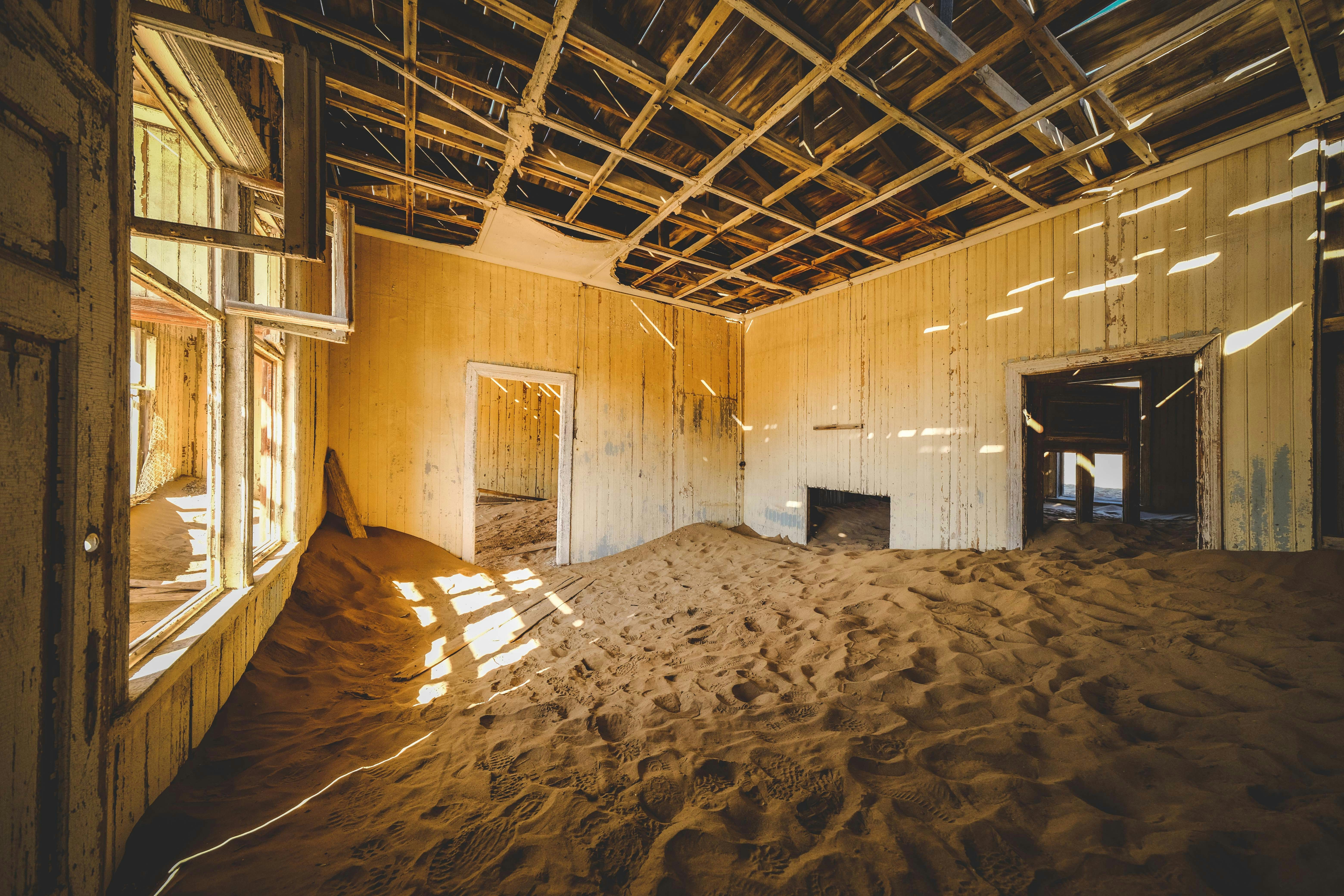 Habitación abandonada con el suelo cubierto de arena, paredes amarillas y techo de madera expuesto.