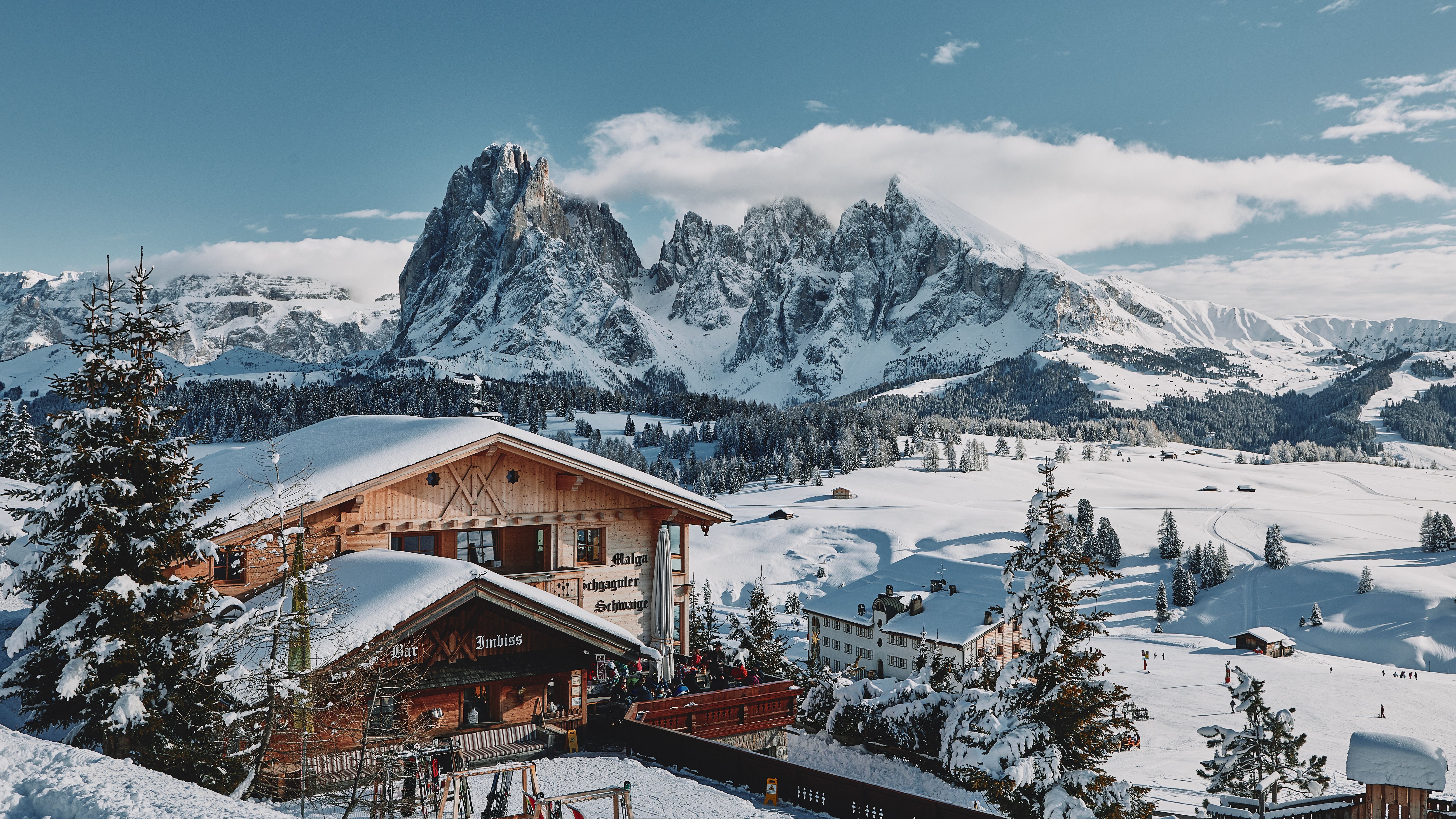 Cabañas de madera cubiertas de nieve con montañas nevadas y pinos bajo un cielo azul.