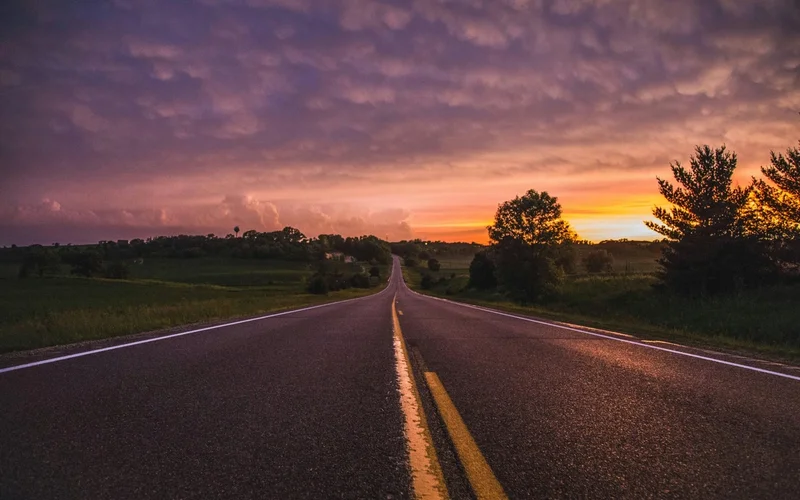 Un camino rural recto se extiende bajo un cielo púrpura tormentoso al atardecer, flanqueado por campos verdes y árboles dispersos