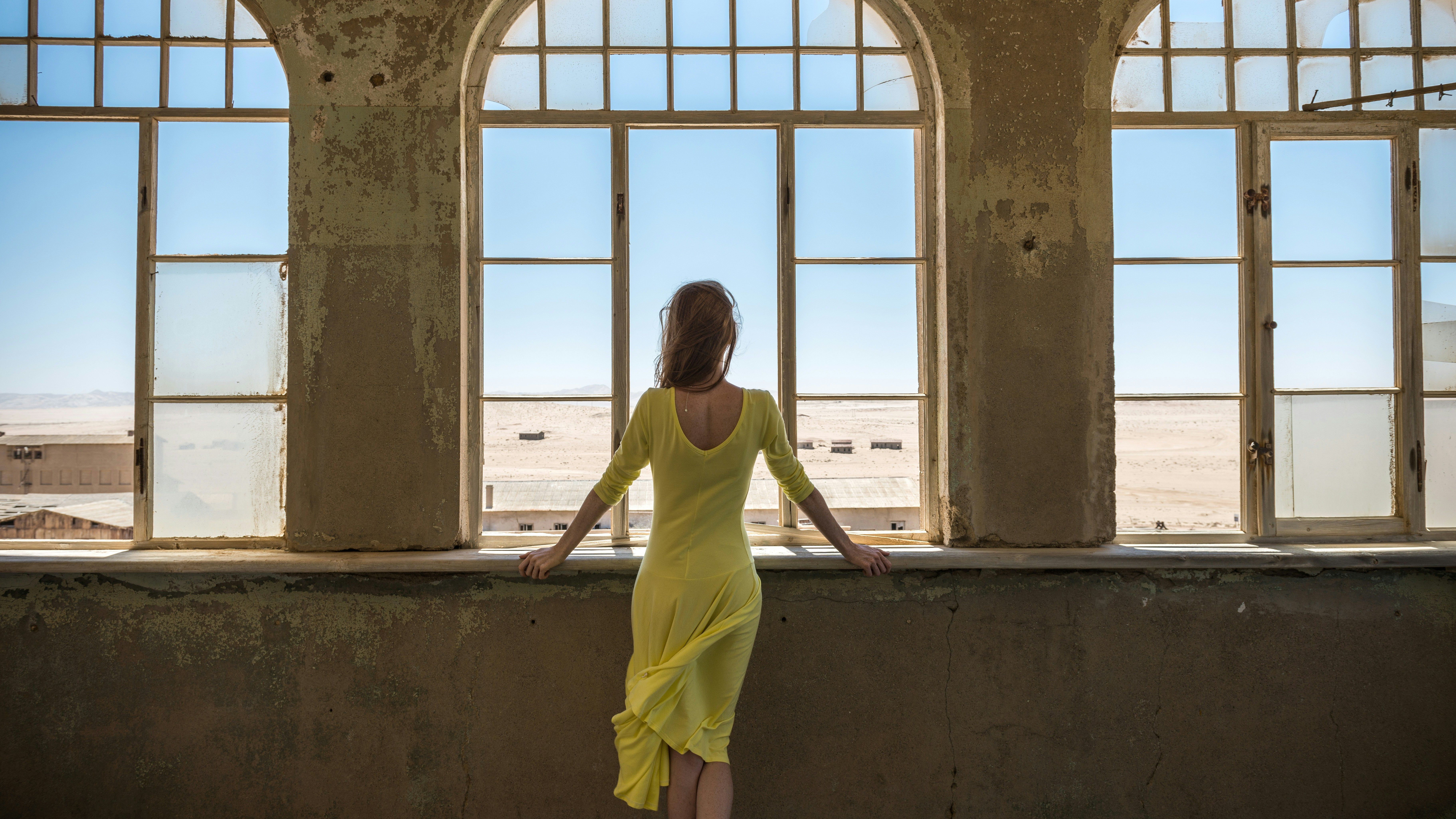 Una persona de espaldas con un vestido amarillo mira a través de grandes ventanas arqueadas hacia un paisaje desértico.