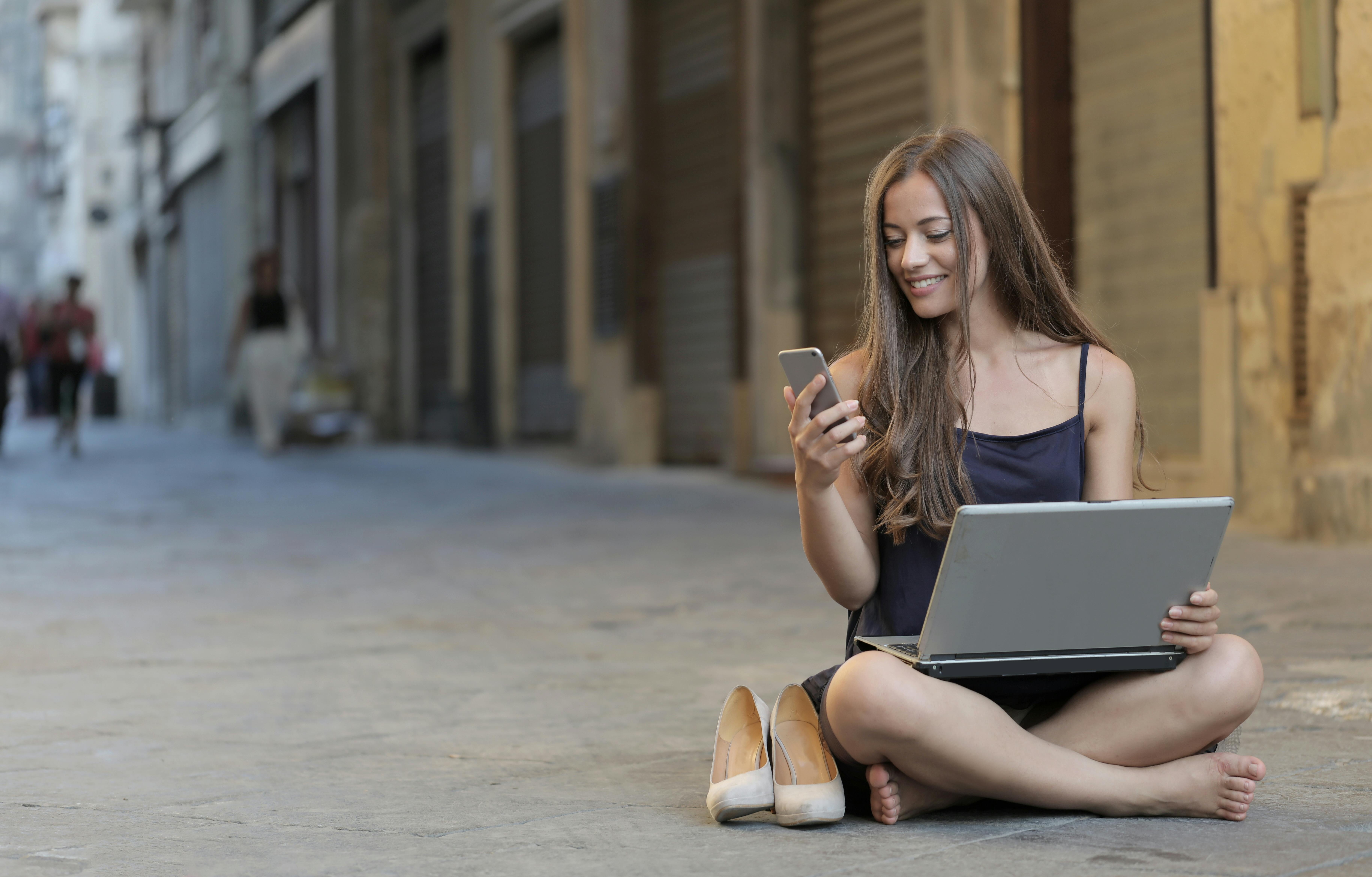 Mujer joven sentada en el suelo de una calle adoquinada, mirando su teléfono con un portátil en el regazo.