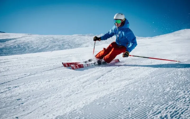 Un esquiador con chaqueta azul y pantalones rojos desciende una pista nevada bajo un cielo azul, rodeado de nieve fresca en las montañas