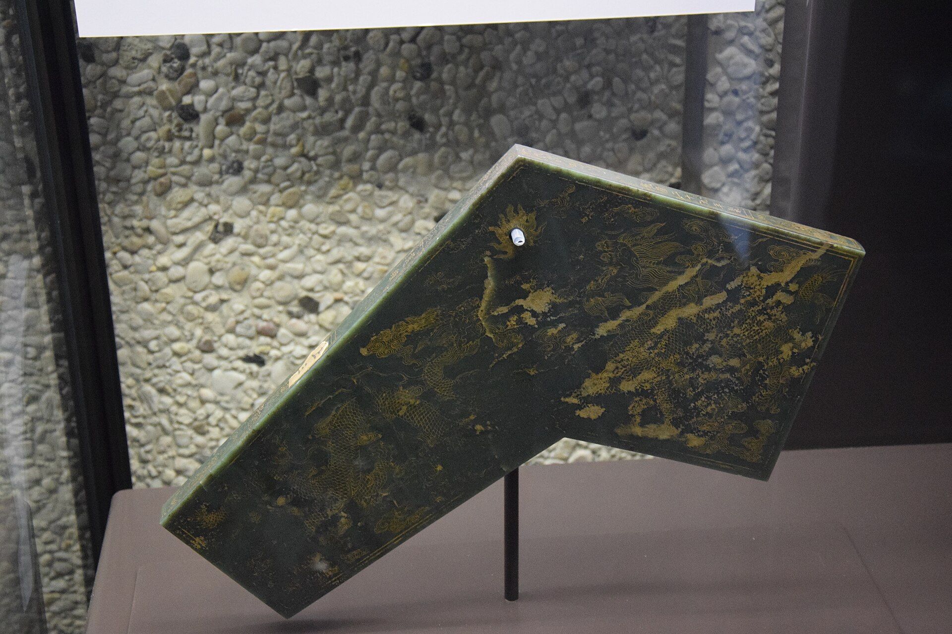 Una losa de jade oscuro con grabados dorados, exhibida en un soporte.