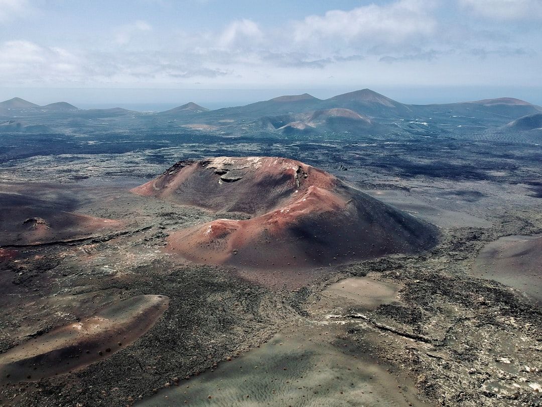Paisaje volcánico de Timanfaya con colinas de lava roja y negra, cráteres y montañas lejanas bajo un cielo azul con nubes