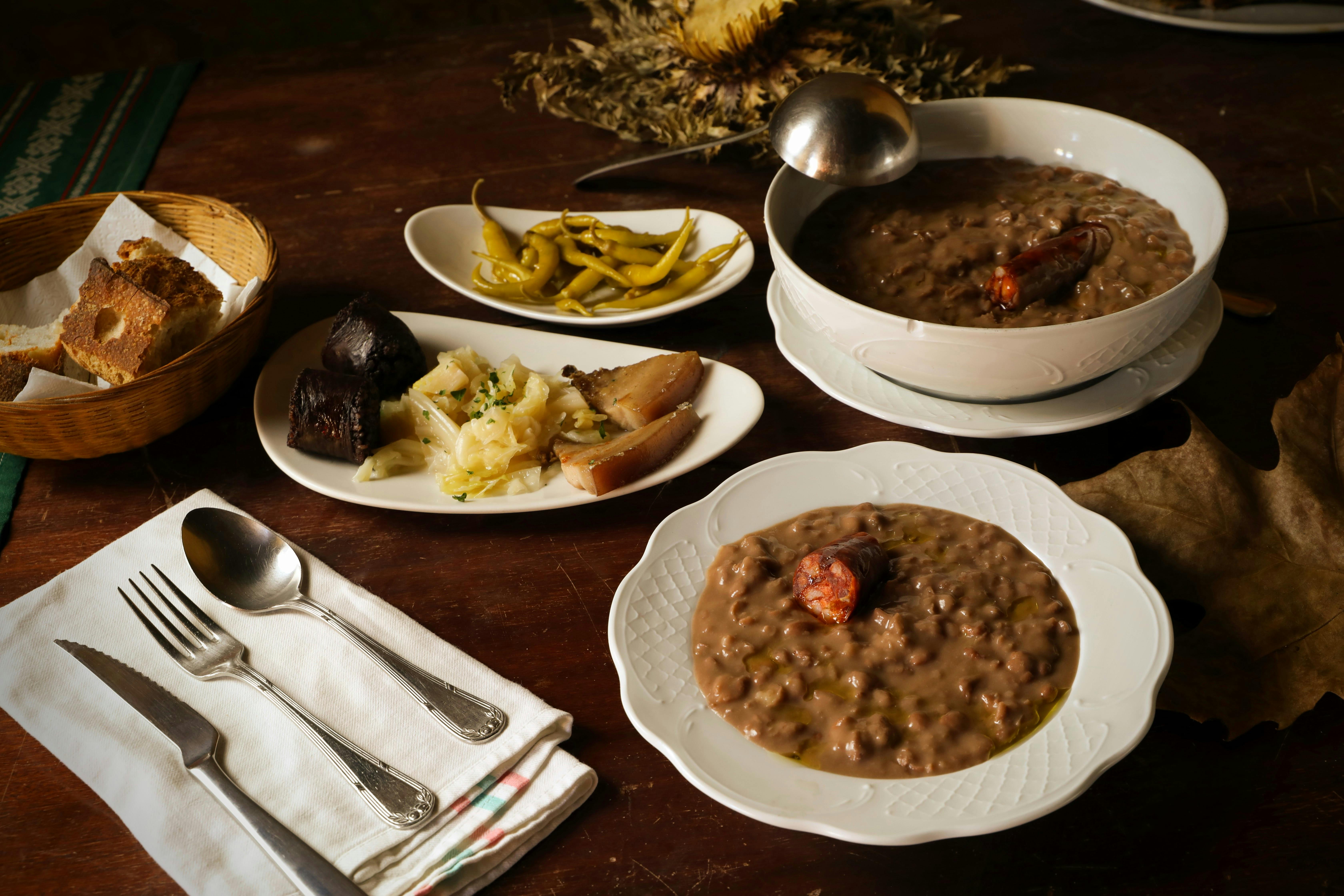 Una mesa de madera con dos platos de legumbres guisadas, pan, pimientos y embutidos.