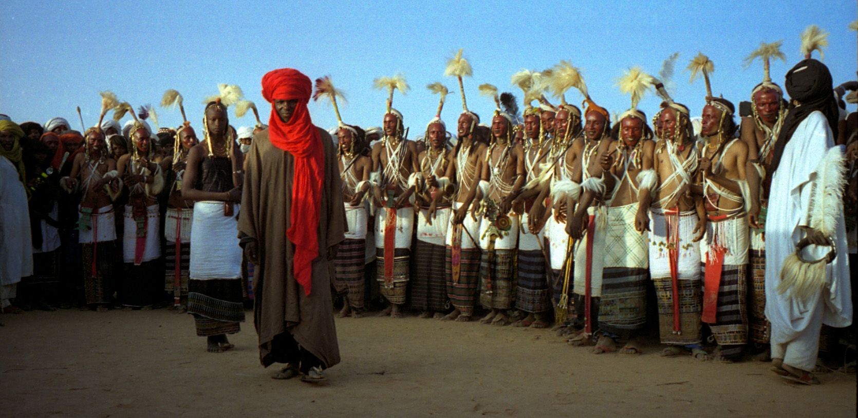 Persona con turbante rojo y túnica parda camina por delante de una larga fila de bailarines con plumas y faldas blancas, sobre la arena.