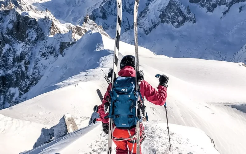 Esquiador de espaldas con chaqueta roja, mochila azul y esquís, en una cresta nevada de montañas rocosas en Chamonix