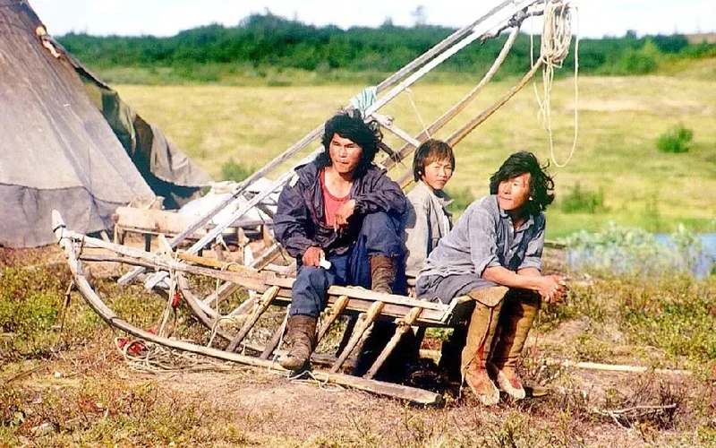 Tres nenets sentados en un trineo de madera en la tundra ártica, con una yurta tradicional al fondo