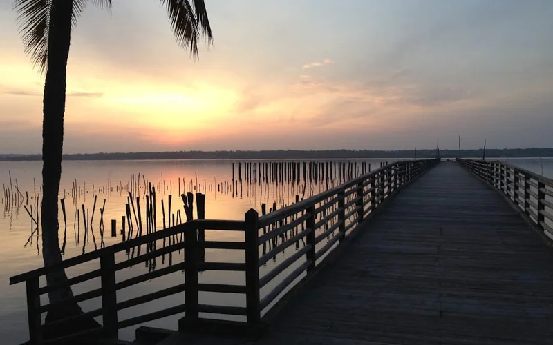 Muelle de madera extendiéndose sobre un lago al atardecer en Benin, con palmeras y postes sumergidos en el agua