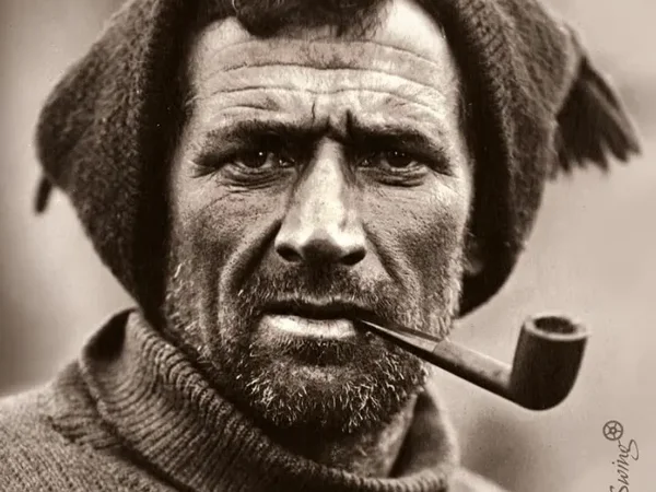 Retrato en blanco y negro de un hombre mayor con barba, fumando una pipa y luciendo gorro de lana y suéter abrigado