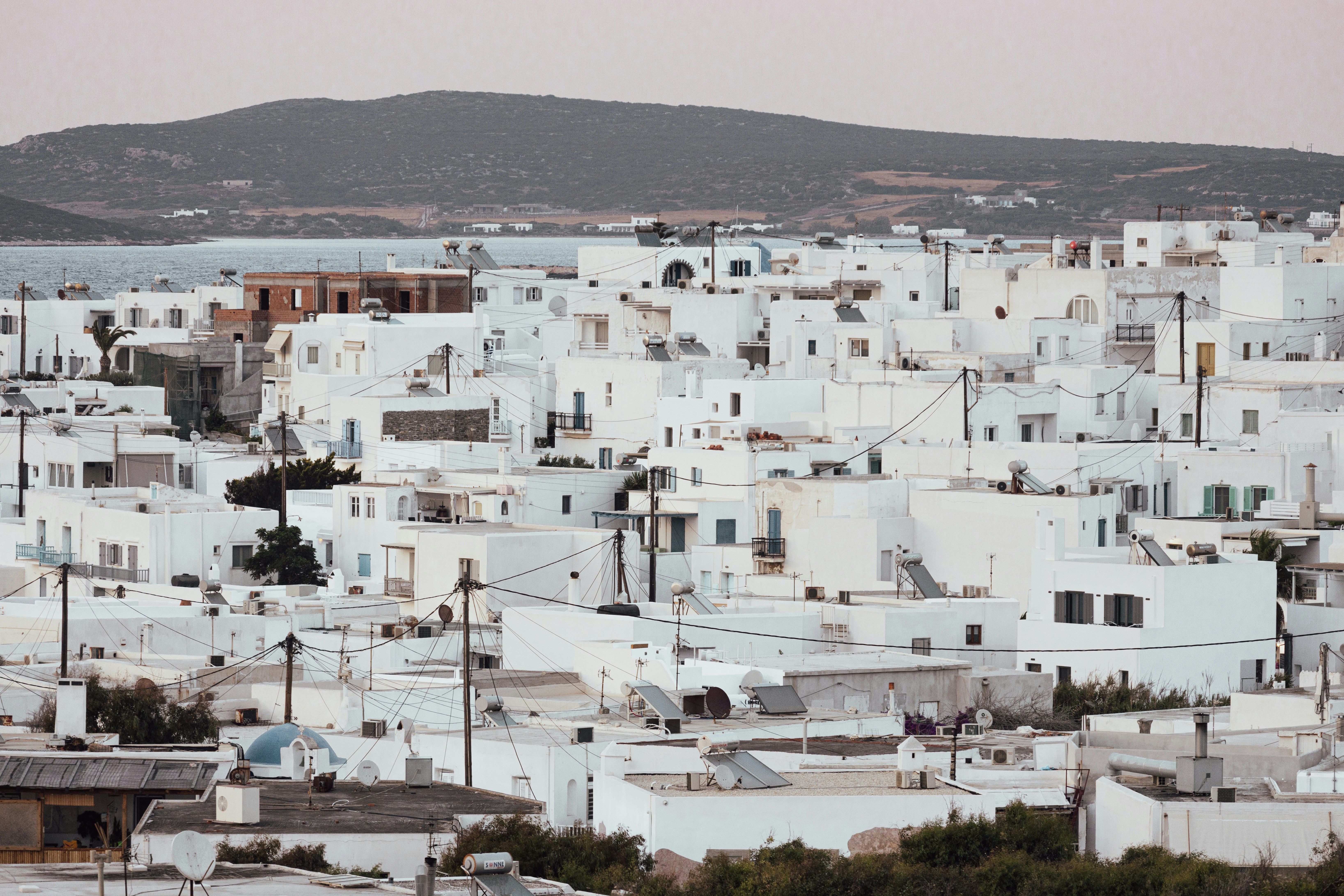 Vista aérea de un pueblo costero con casas blancas y tejados planos, con el mar y montañas al fondo.